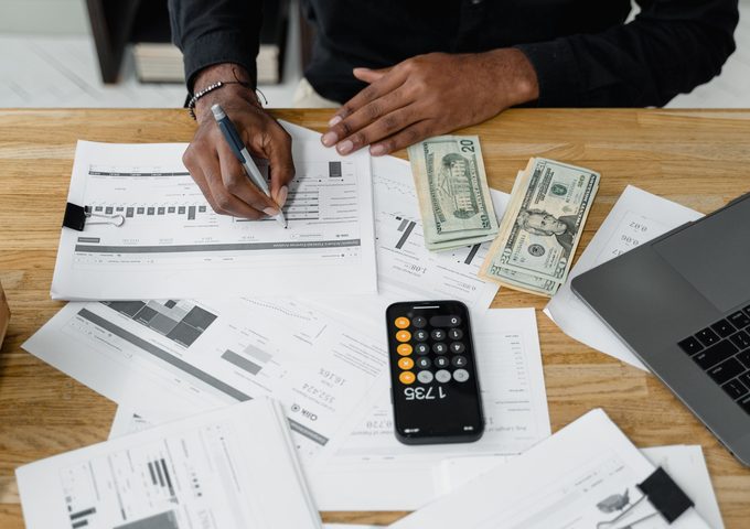 Hands writing calculations on financial documents beside a calculator, cash, and a laptop